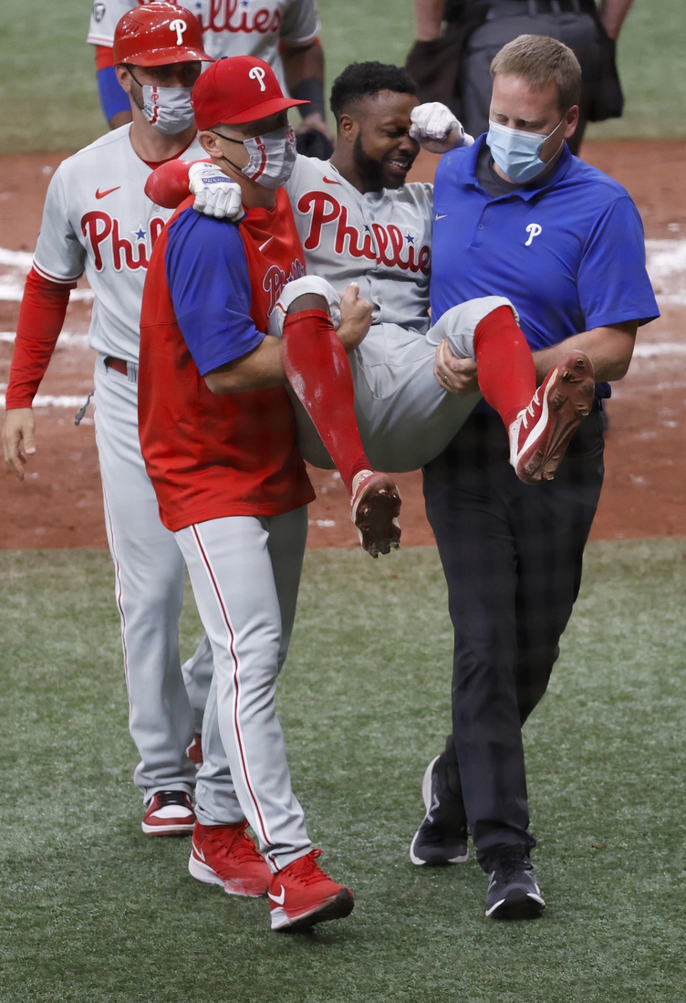 May 29, 2021; St. Petersburg, Florida, USA; Philadelphia Phillies center fielder Roman Quinn (24) is carried off the field by Philadelphia Phillies manager Joe Girardi (25) and trainer after an apparent injury as he scores s run during the fifth inning against the Tampa Bay Rays  at Tropicana Field. Mandatory Credit: Kim Klement-USA TODAY Sports