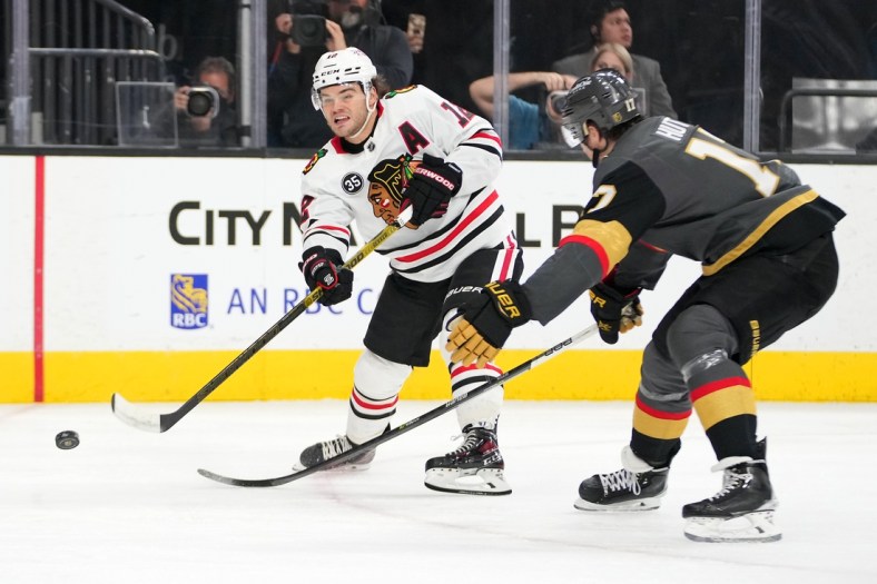 Mar 26, 2022; Las Vegas, Nevada, USA; Chicago Blackhawks left wing Alex DeBrincat (12) shoots in front of Vegas Golden Knights defenseman Ben Hutton (17) during the first period at T-Mobile Arena. Mandatory Credit: Stephen R. Sylvanie-USA TODAY Sports