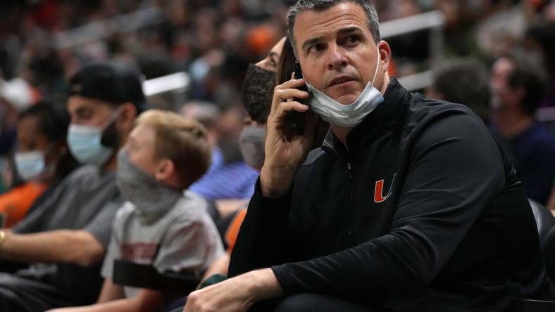 Feb 2, 2022; Coral Gables, Florida, USA; Miami Hurricanes head football coach Mario Cristobal talks on a phone while sitting court-side during the second half between the Miami Hurricanes and the Notre Dame Fighting Irish at Watsco Center. Mandatory Credit: Jasen Vinlove-USA TODAY Sports