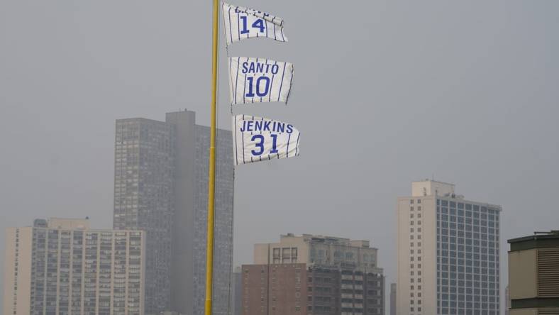 Jun 27, 2023; Chicago, Illinois, USA; The flags of Chicago Cubs' Hall of Fame players Ernie Banks (14), Ron Santo (10) and Ferguson Jenkins fly off the Wrigley Field left field foul pole as a veil of haze from Canadian wildfires shroud high rise buildings along Lake Michigan before a baseball game between the Cubs and the Philadelphia Phillies at Wrigley Field. Mandatory Credit: David Banks-USA TODAY Sports