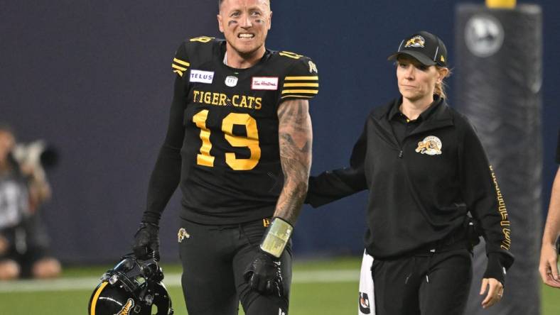 Jun 18, 2023; Toronto, Ontario, CAN;  Hamilton Tiger-Cats quarterback Bo Levi Mitchell (19) is escorted off the field by  team head therapist Claire Tofflemire after suffering an injury against the Toronto Argonauts in the fourth quarter at BMO Field. Mandatory Credit: Dan Hamilton-USA TODAY Sports