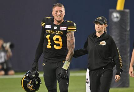 Jun 18, 2023; Toronto, Ontario, CAN;  Hamilton Tiger-Cats quarterback Bo Levi Mitchell (19) is escorted off the field by  team head therapist Claire Tofflemire after suffering an injury against the Toronto Argonauts in the fourth quarter at BMO Field. Mandatory Credit: Dan Hamilton-USA TODAY Sports