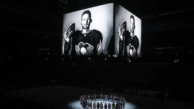 Oct 30, 2023; Pittsburgh, Pennsylvania, USA;  Members of the Anaheim Ducks and the Pittsburgh Penguins stand at center ice for a moment of silence at the PPG Paints Arena to honor former Penguin forward Adam Johnson (on scoreboard) who was tragically killed in a hockey related accident in Europe. Mandatory Credit: Charles LeClaire-USA TODAY Sports