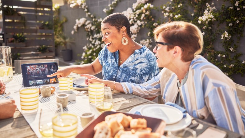 Two Girlfriends Watching Sling TV On Their Tablet While Eating On Their Patio